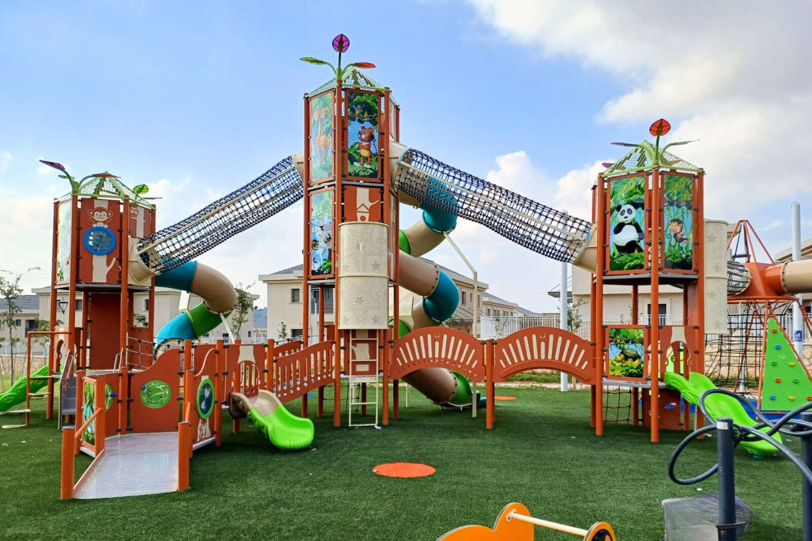 Large themed commercial playground structure with tube slides, net climbing bridges and animal-themed panels on artificial turf